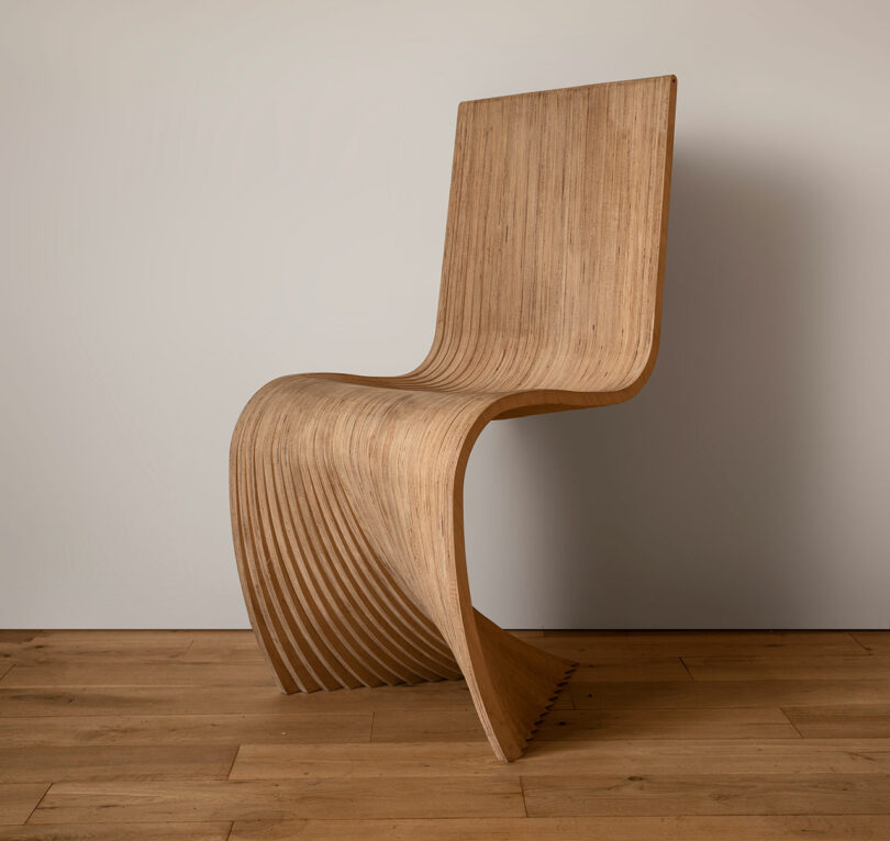 A modern wooden chair with a curvilinear design, featuring a single continuous piece of wood forming the backrest, seat, and legs, placed on a wooden floor against a plain wall.