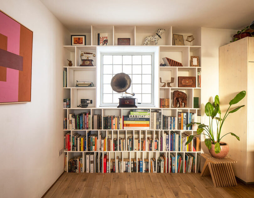 Bookshelf with various objects including books, a gramophone, and decorations, positioned around a window. A large potted plant is placed on the right, and a colorful painting hangs on the left wall.