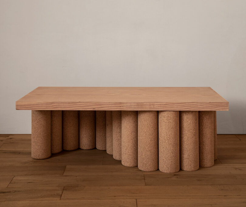 A rectangular wooden table with cylindrical cork legs arranged in a semi-circle pattern, set on a wooden floor against a plain wall.