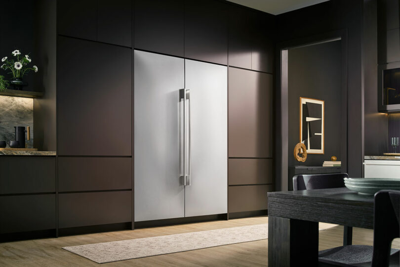 Modern kitchen with dark cabinets, a built-in Signature Kitchen Suite stainless steel refrigerator, minimalist decor, and a light-colored rug on wooden flooring.