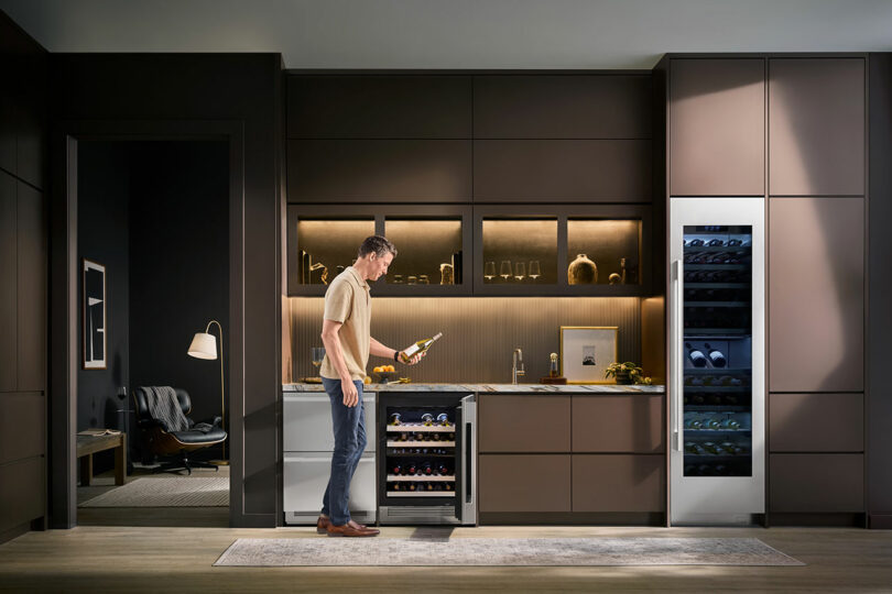 A man stands in a modern kitchen, holding a wine bottle and examining a wine refrigerator in the Signature Kitchen Suite, surrounded by cabinets and appliances.