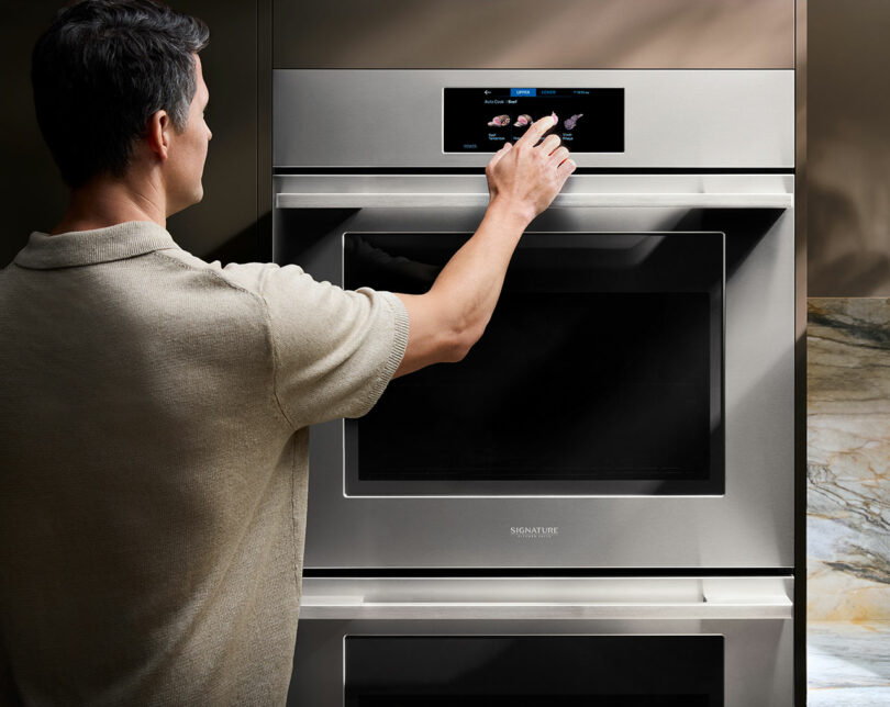 A person adjusts settings on a Signature Kitchen Suite stainless steel wall oven with a digital touchscreen control panel.