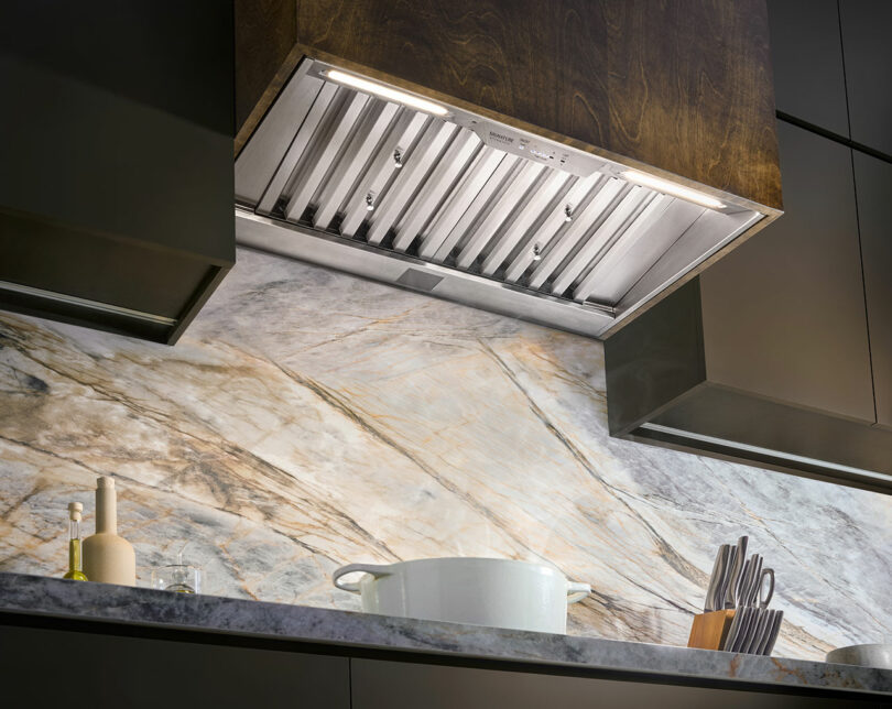 Modern kitchen with a Signature Kitchen Suite built-in stainless steel range hood, marble backsplash, and a countertop featuring a white pot, knife set, and kitchen utensils.