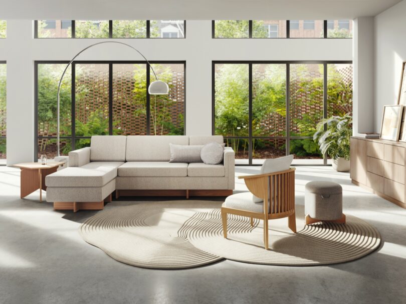 Modern living room with large windows, a cream-colored sectional sofa, a wooden chair, a matching ottoman, a floor lamp, and a round beige rug on a gray concrete floor. Channeling Frank Lloyd Wright's design ethos, the outdoor large plants add an organic touch to the space.