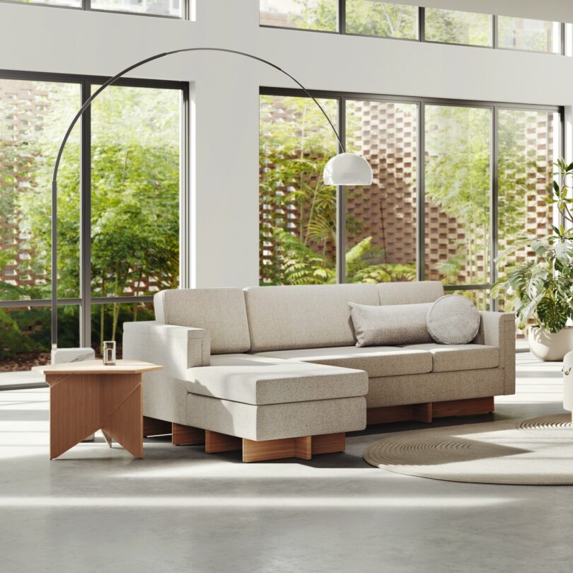 A modern living room, inspired by Frank Lloyd Wright, features a beige sectional sofa, a wooden side table, a curved floor lamp, and large windows overlooking a lush garden.