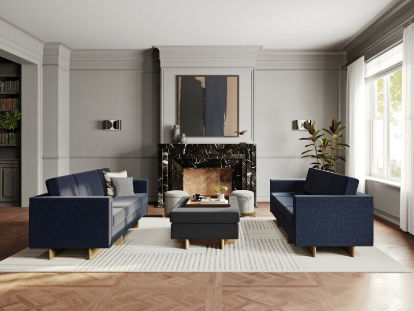 A modern living room inspired by Frank Lloyd Wright features two navy blue sofas, a black marble fireplace, a central black ottoman, a potted plant, and shelving units, with large windows allowing natural light to enter.