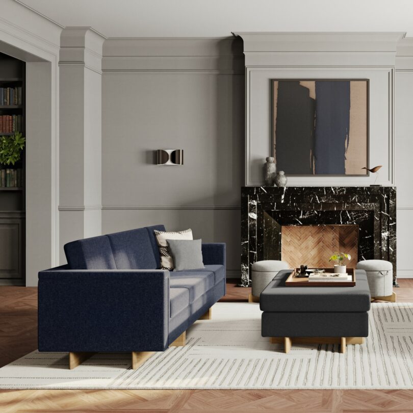 A modern living room with a blue sofa, matching ottoman, marble fireplace, abstract artwork, and grey walls showcases design elements that echo the innovative spirit of Frank Lloyd Wright.