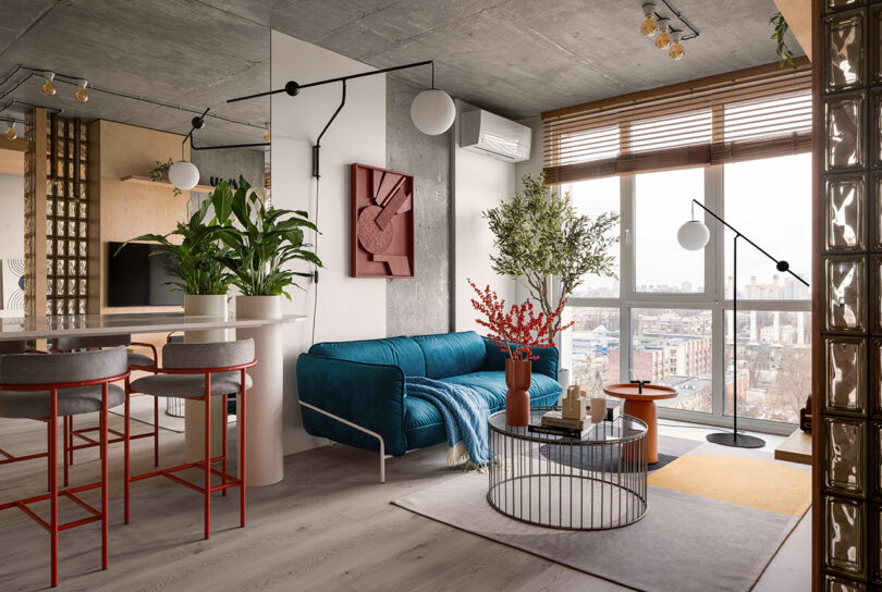 Modern living room with blue sofa, round coffee table, houseplants and large windows overlooking the cityscape. Featuring industrial style elements and contemporary decor.