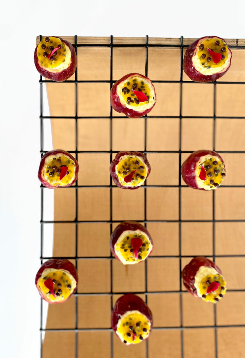 Small yellow and red passion fruits topped with seeds on a cooling rack over a wooden surface, arranged in a grid pattern.