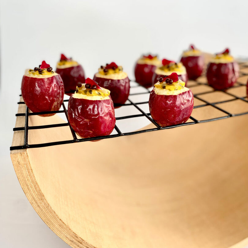 Red-colored, round pastries filled with a yellow cream and garnished with red and black toppings are arranged on a black wire rack. The wire rack is placed on a curved wooden surface, adding an extra layer of elegance to the display that subtly hints at exotic ingredients like coconut tapioca.