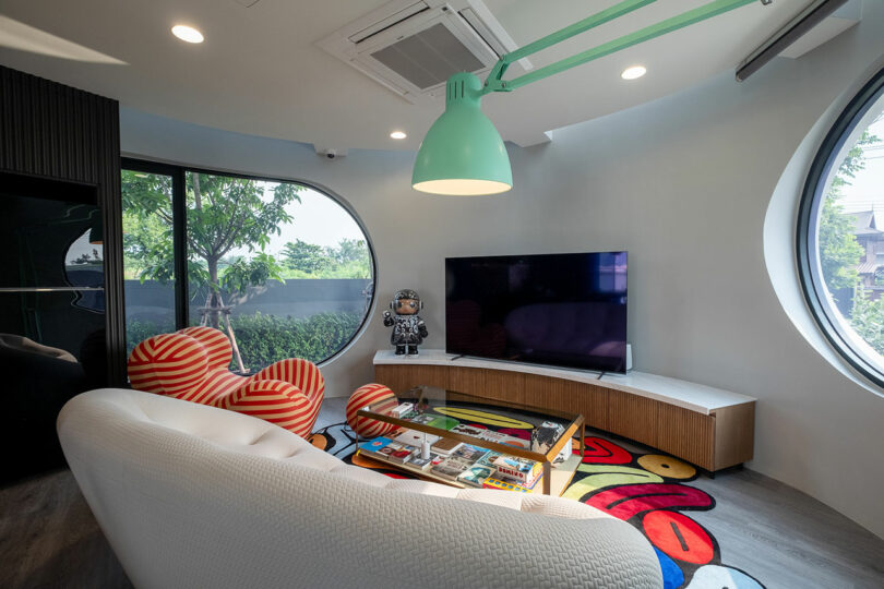 A modern room with circular windows features a large TV, a white sofa, colorful seating, a coffee table with magazines, and a green overhanging lamp. A decorative figure stands near the TV.