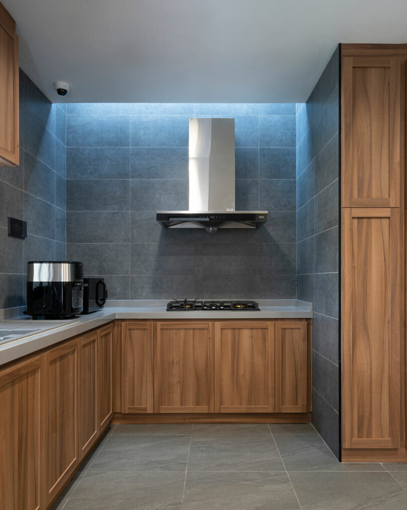 Modern kitchen with wooden cabinets, stainless steel range hood, built-in stovetop, coffee maker, and dark gray tiled walls and floor.