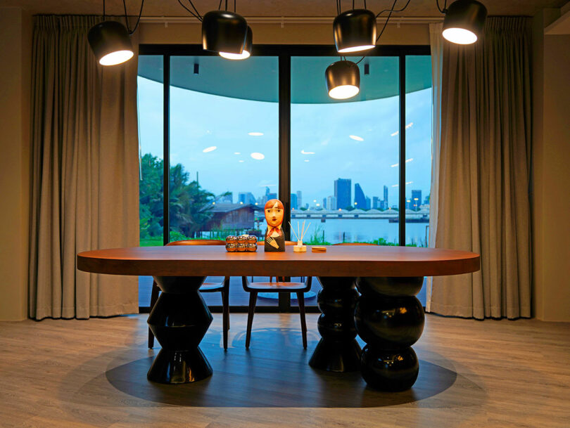 A modern dining area features a large table with unique, sculptural legs, and a city skyline visible through expansive windows in the background.