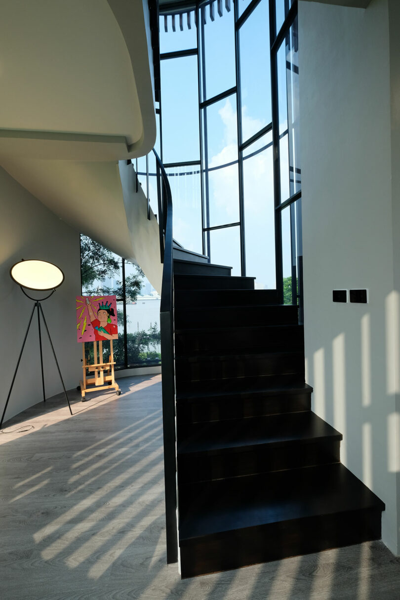 A modern interior with a black staircase, a tripod floor lamp, and an easel supporting a colorful painting, all illuminated by large, curved floor-to-ceiling windows.