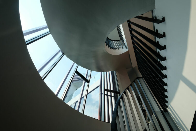 A view looking up a spiral staircase with metal railings and large windows allowing natural light to illuminate the interior.