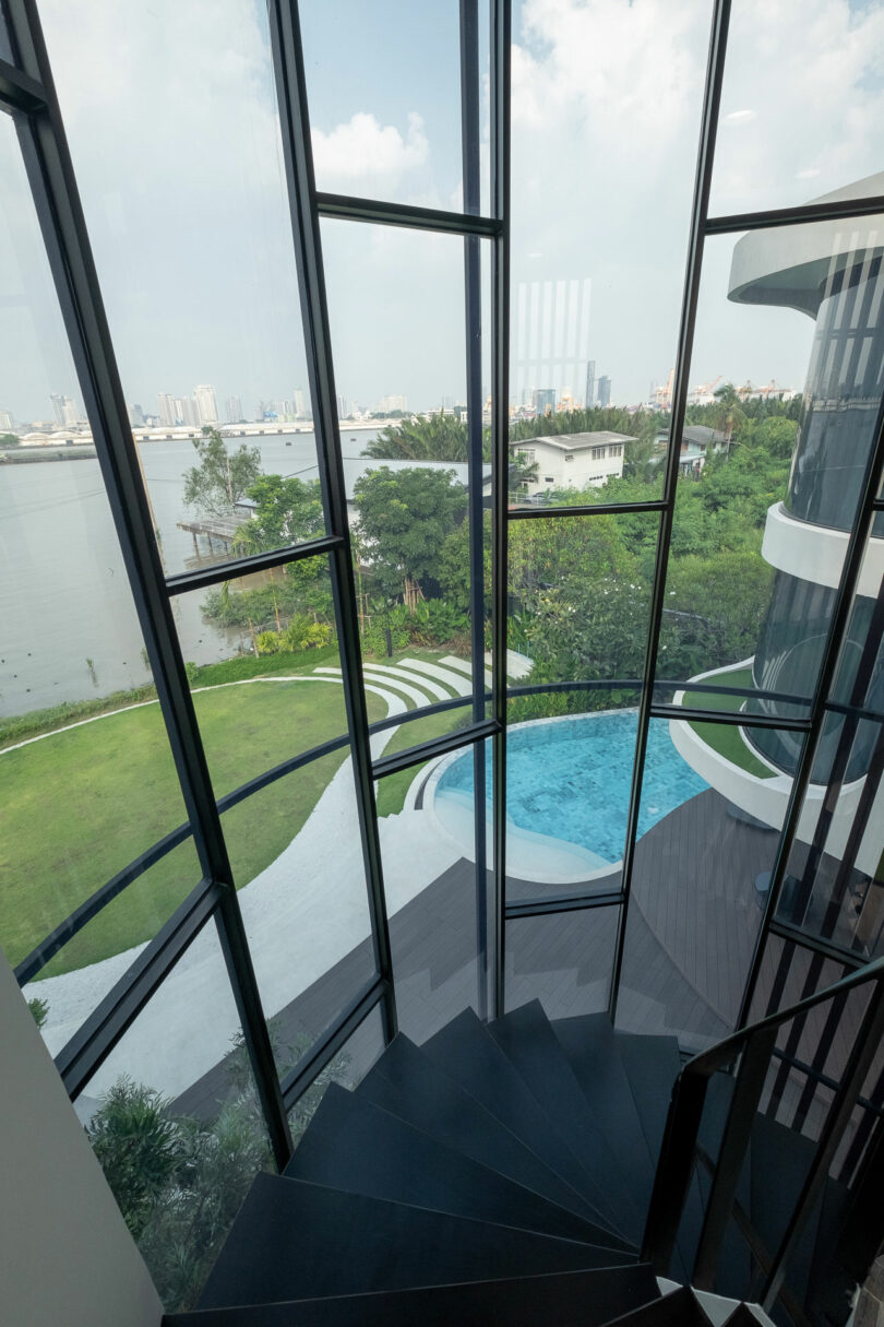 View from a spiral staircase landing, looking out through large windows at a curved swimming pool, green lawn, and distant city skyline across a body of water.