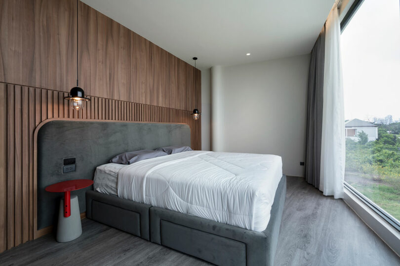 A modern bedroom featuring a double bed with a gray upholstered frame, a wooden accent wall, hanging pendant lights, and a red side table, all adjacent to a large window with a view of greenery.