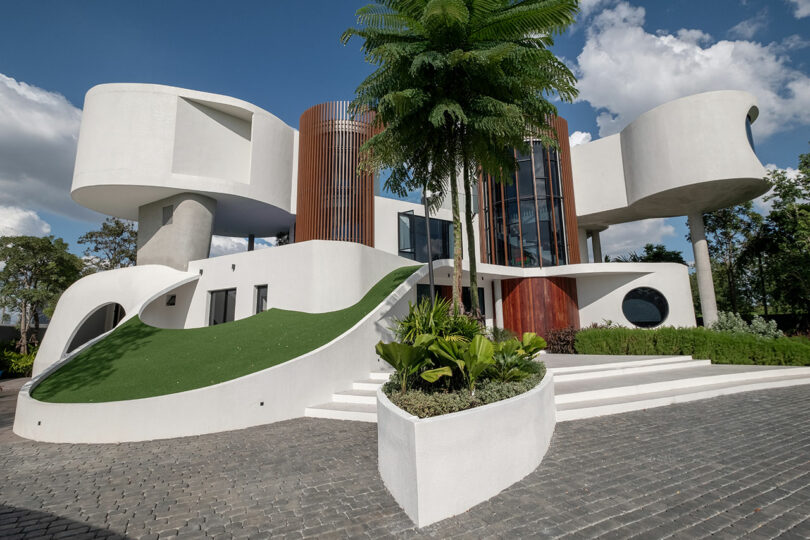 A modern building with a unique, curvy design features white exterior, large windows, and green landscaping on a sunny day.