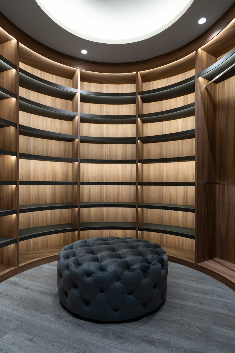 A circular wooden shelving unit with empty shelves and a round, tufted ottoman in the center. The room is illuminated by a circular overhead light.