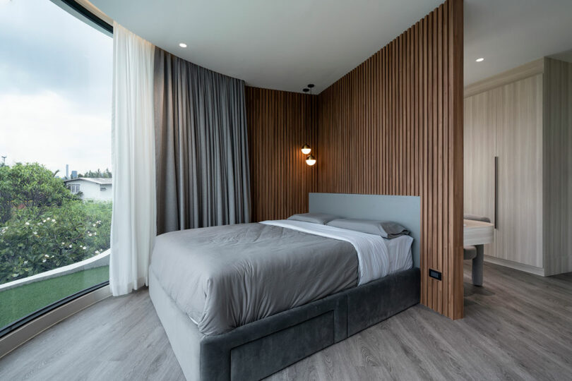 A modern bedroom with a double bed, wood paneling on one wall, a curved window with sheer and blackout curtains, and adjacent bathroom. The room has gray bedding and light wood flooring.