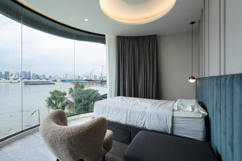 A modern bedroom features a curved floor-to-ceiling window with a cityscape view, a bed with white linens, a gray armchair, pendant light, and gray curtains.