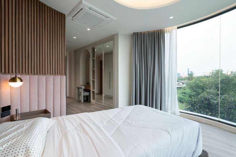 A modern bedroom with large floor-to-ceiling windows, a bed with white bedding, a padded headboard, a wall-mounted lamp, a wooden wall panel, and a dressing area with mirrors.