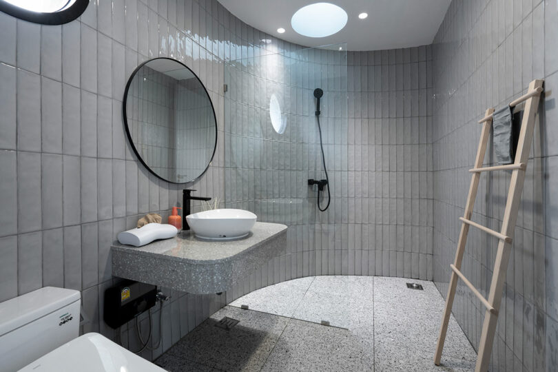 Modern bathroom with gray tiled walls, a round mirror above a floating sink, a walk-in shower, a toilet, and a wooden ladder-style towel rack. Circular skylight and recessed lights provide illumination.