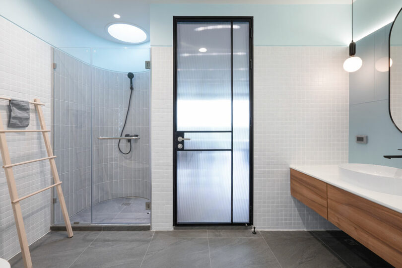 Modern bathroom featuring a glass-enclosed shower, a frosted glass door with black frame, a ladder towel rack, a wooden vanity with dual sinks, and a round mirror.