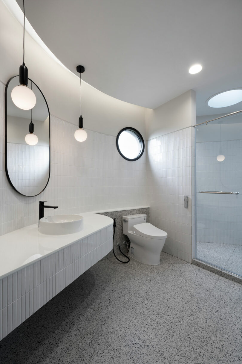 Modern bathroom with a minimalist design, featuring a wall-mounted oval mirror, two hanging light fixtures, a white countertop with a sink, a toilet, and a walk-in shower with glass doors.