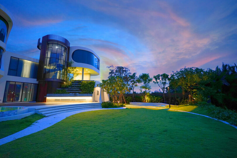 Modern house with expansive, well-manicured lawn and trees at dusk, featuring large windows, curved architectural elements, and outdoor lighting.