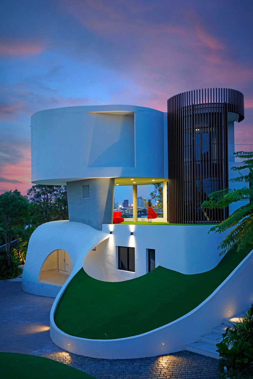 A modern, curved white house with large windows, green lawns, and a circular balcony lit by ambient lighting against a vivid dusk sky.