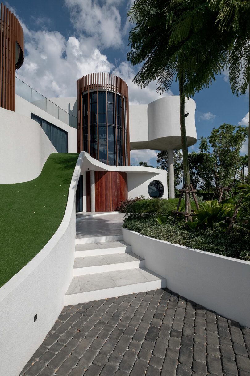 Modern building with a curved white facade, featuring a wooden door, circular windows, and a glass cylindrical tower. The entrance path has steps and greenery, with a large tree in the foreground.