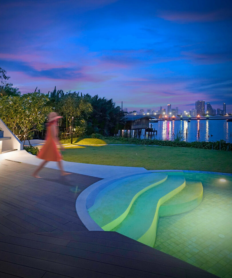 A person in a red dress walks near a modern pool with curved steps at dusk, overlooking a river and a city's illuminated skyline in the distance. The scene includes trees and distant buildings.