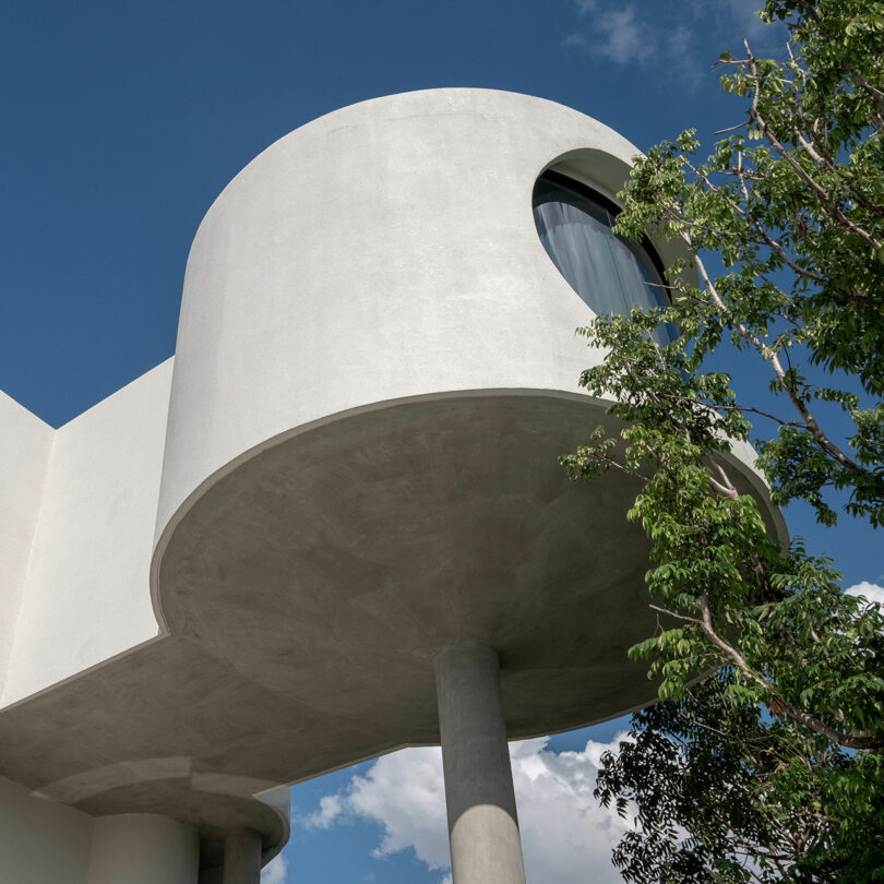 A modern white building with a cylindrical upper floor supported by columns and featuring a large round window. A tree partially obscures part of the building, set against a blue sky with some clouds.