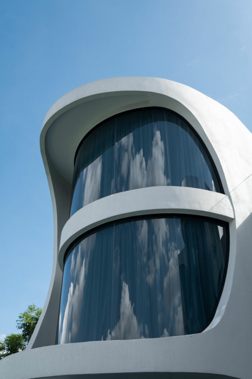 Modern white building with two rounded, curved glass windows reflecting clouds and blue sky, set against a clear blue background. Green foliage can be seen at the building's base.