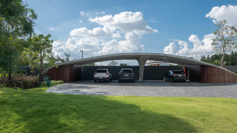 Modern carport with a curved roof design, housing three parked cars on a paved driveway with a grassy lawn in the foreground and a clear blue sky with scattered clouds in the background.