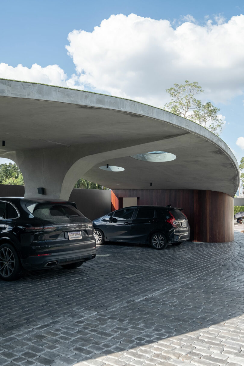 Two black cars are parked under a modern, curved concrete carport with a cobblestone driveway. The structure has circular skylights and is surrounded by trees and a partly cloudy sky.