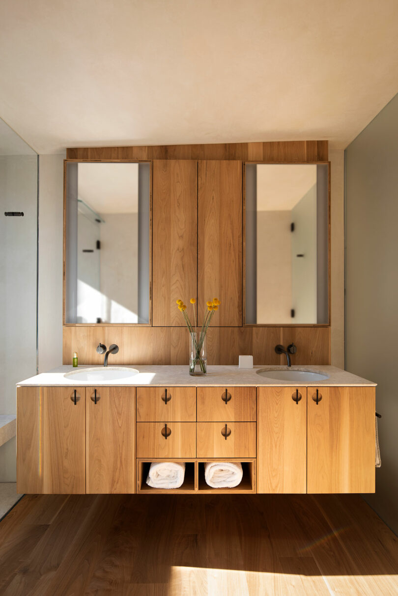 Modern bathroom with a wooden double-sink vanity, two mirrors, a central vase with yellow flowers, and neatly folded towels on the lower shelves.