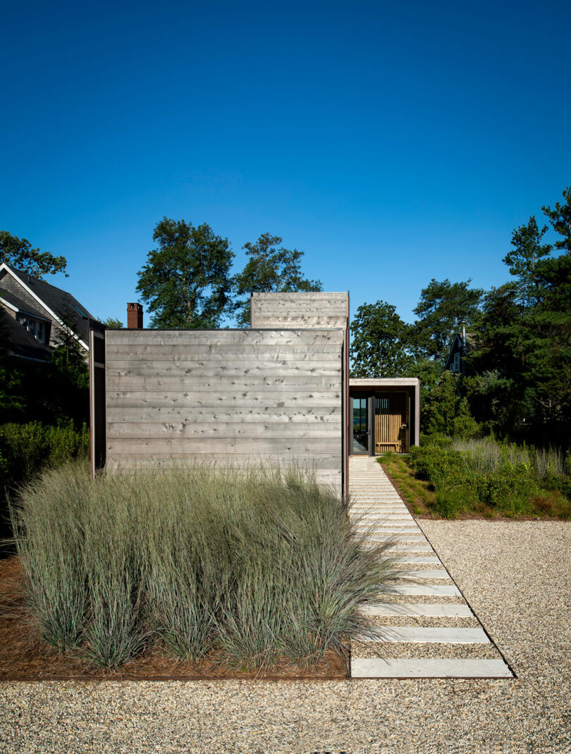 A modern, minimalist house with wooden exterior walls and a stone pathway leading to the entrance, surrounded by tall grass and trees, under a clear blue sky.