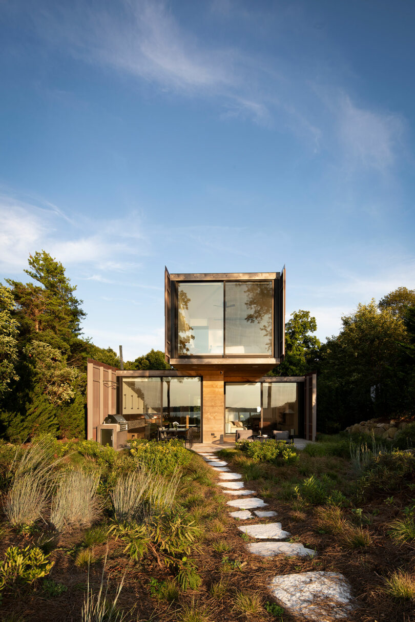 A modern, two-story house with large glass windows and a wooden exterior stands amidst a garden with a stone pathway leading up to it. The sky is clear and blue.