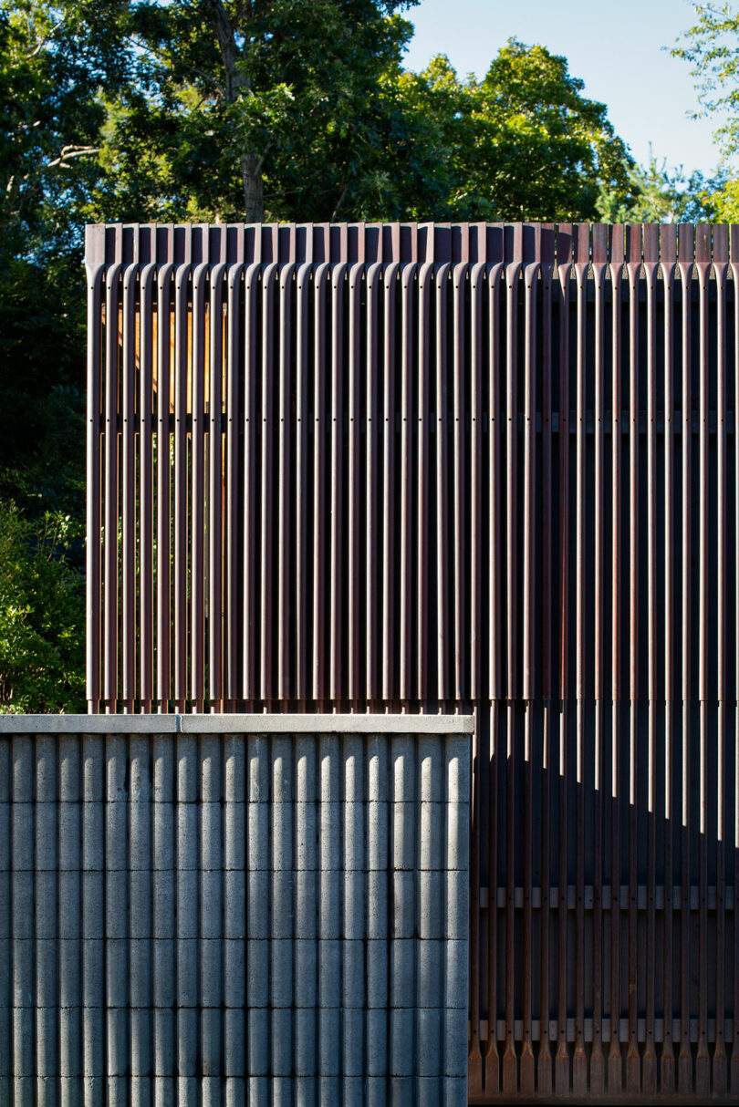 Close-up of a modern building facade featuring vertical wooden slats on the upper section and horizontal concrete blocks on the lower section, with trees visible in the background.