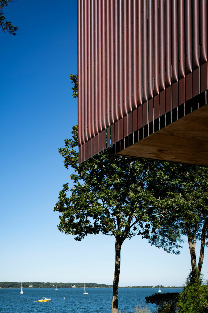 Close-up view of a building with red vertical panels and a clear blue sky background, overlooking a body of water with sailboats and trees.