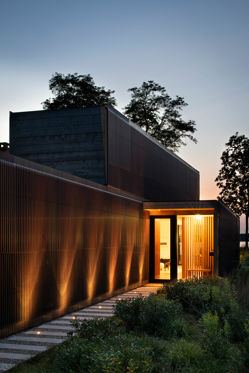 A modern building exterior at dusk, featuring sleek lines and vertical wooden slats, with warm interior lighting visible through large windows. Trees are seen in the background.