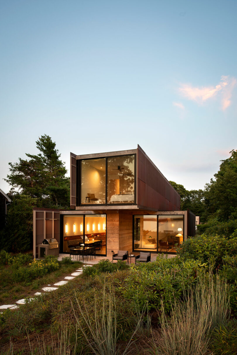 Modern two-story house with large glass windows, surrounded by greenery. A patio with outdoor seating is visible in the foreground.