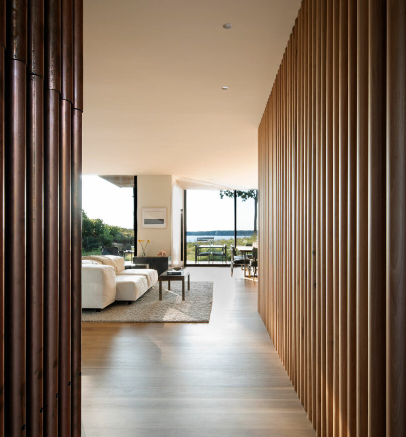 A modern, minimalist living room with light wood flooring, white sofas, a wooden slat wall divider, and large windows overlooking an outdoor landscape.