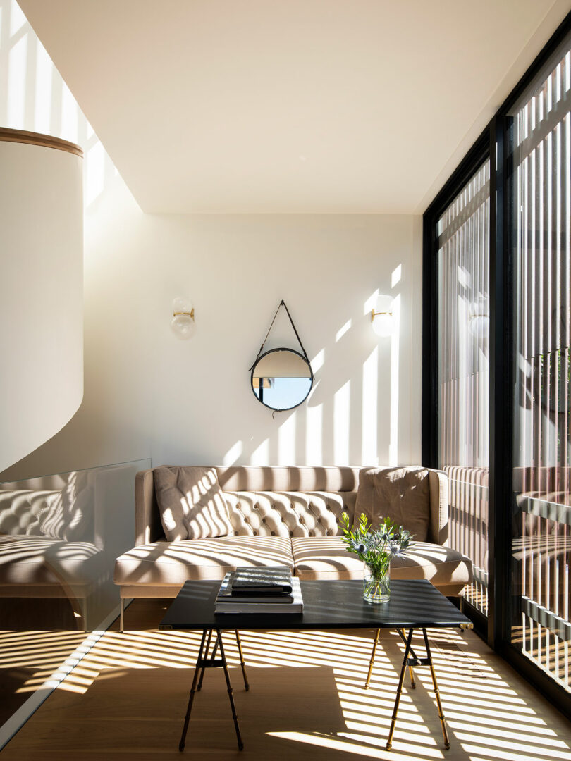 A well-lit modern living room with a beige sofa, round mirror, black coffee table, and large windows covered by vertical blinds casting shadows. A small plant and books are on the table.