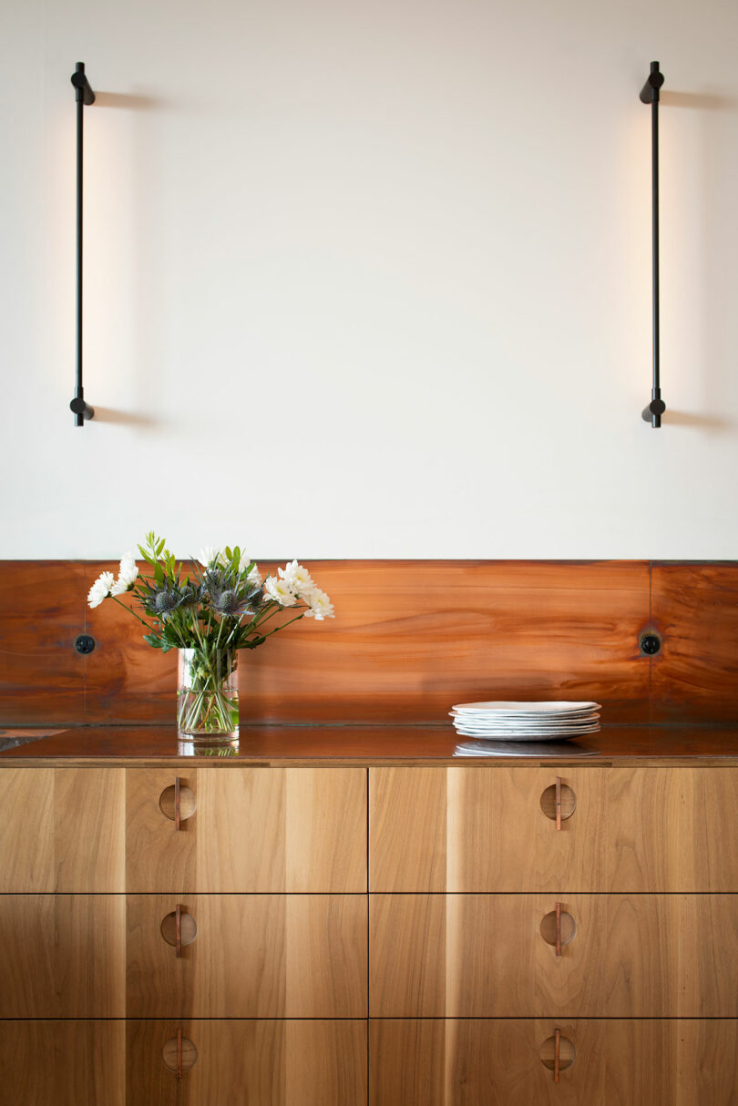 A kitchen with wooden cabinetry, a glass vase with flowers, a stack of plates on the counter, and two wall-mounted light fixtures.