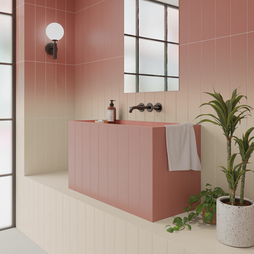 Modern bathroom with gradient pink tiles, a rectangular pink bathtub, black fixtures, a wall-mounted light, and a large window. A potted plant and towel are placed on the tub's ledge.