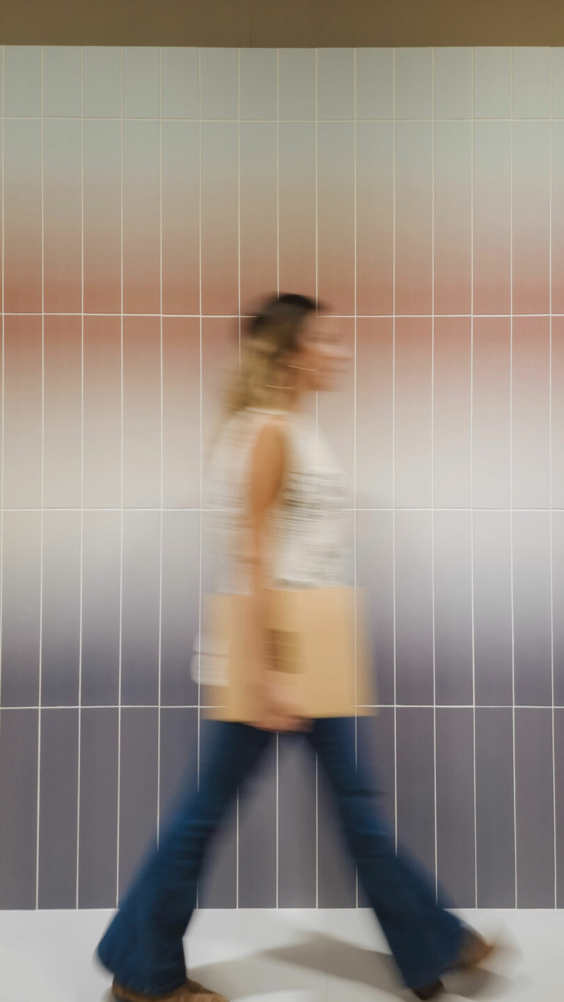 Blurred image of a person walking in front of a gradient-colored tiled wall, holding a folder.