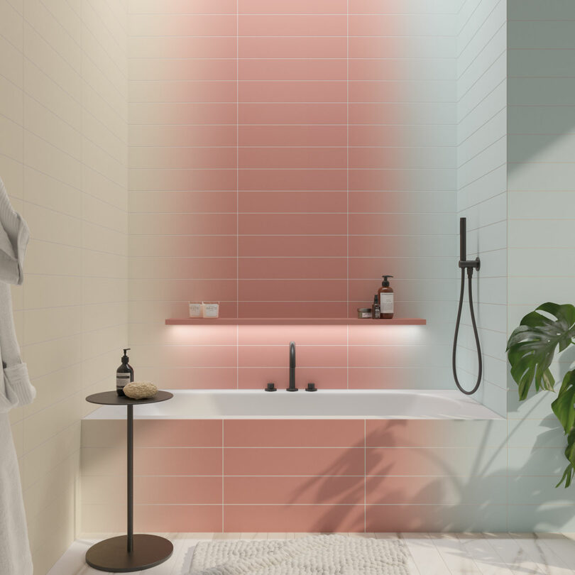 Modern bathroom with gradient pink and white tiles, a built-in bathtub, black faucet, handheld showerhead, and a round side table holding bath items. A leafy plant and wall-mounted shelf are also present.